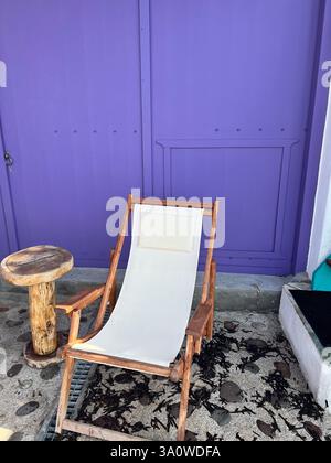 Una sedia a sdraio in legno con un'imbragatura bianca si trova accanto a un'audace parete viola Foto Stock