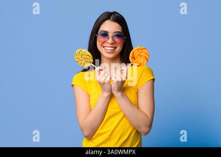 Dolcezza. Bella giovane donna estate che copre l'occhio con grande luminoso lollipop e guardando la macchina fotografica, sfondo giallo con spazio di copia Foto Stock
