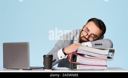 Uomo d'affari sopraffaricato sdraiato sulla scrivania vicino al computer portatile su sfondo blu, spazio vuoto Foto Stock
