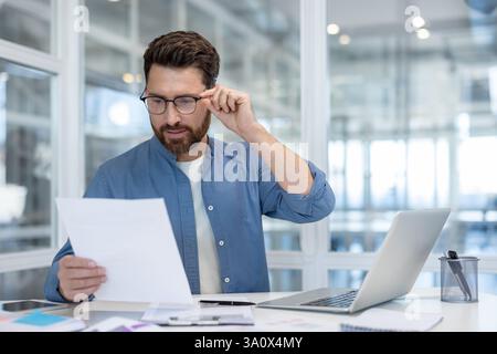 Un uomo con la barba esamina i documenti aziendali in un ufficio luminoso, utilizzando un notebook. Sta indossando occhiali e una camicia blu. Foto Stock