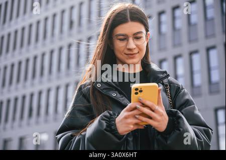 Una giovane donna è all'aperto vicino all'edificio, è in piedi e tiene in mano lo smartphone. Foto Stock
