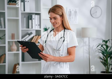 Lettura delle informazioni nel blocco note. Giovane medico donna in camice bianco è al chiuso. Foto Stock