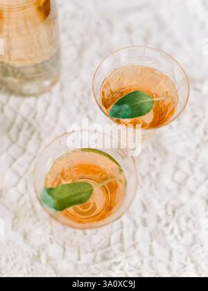 Due bicchieri di cocktail scintillanti adornati con foglie di menta fresche si siedono su una delicata tovaglia di pizzo alla luce del giorno. Foto Stock