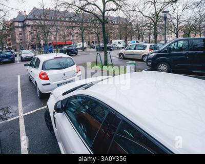 Strasburgo, Francia - 11 febbraio 2025: Parcheggio urbano coperto con diverse auto parcheggiate vicino a edifici storici, alberi senza foglie che si riflettono su marciapiedi bagnati Foto Stock