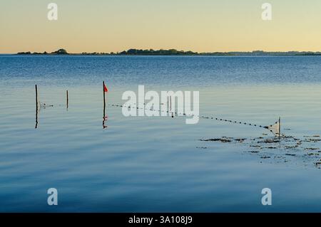Onde dolci si infrangono su una tranquilla zona di pesca segnata da pali e reti, con luce solare calda che illumina la tranquilla scena al crepuscolo. Foto Stock