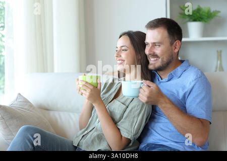 Coppia felice che guarda attraverso una finestra e beve un caffè seduto sul divano di casa Foto Stock