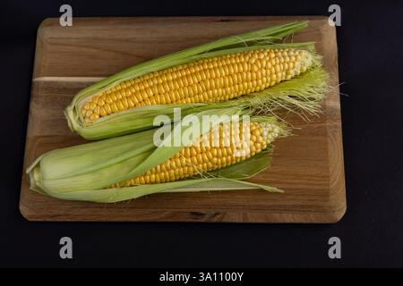 Due orecchie fresche di mais con bucce verdi parzialmente sbucciate, rivelando chicchi dorati gialli, poste su un tagliere di legno contro Foto Stock