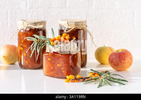 Tre vasetti di spinoso marino e marmellata di mele sono esposti su un tavolo accanto alle mele fresche e ai frutti di spinone marino. Foto Stock