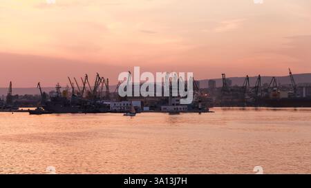Scena portuale di Varna durante l'alba con gru industriali sagomate contro il cielo vibrante, che si specchia sull'acqua calma. Una tranquilla miscela di urban in Foto Stock