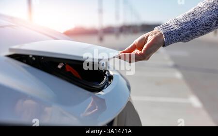 Una persona che indossa un maglione grigio che apre il coperchio della porta di ricarica di un'auto elettrica bianca su una superficie di cemento. Foto Stock