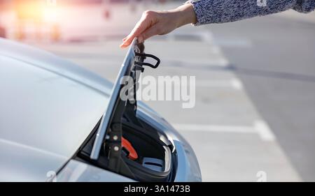 Una persona che indossa un maglione grigio che apre il coperchio della porta di ricarica di un'auto elettrica bianca su una superficie di cemento. Foto Stock