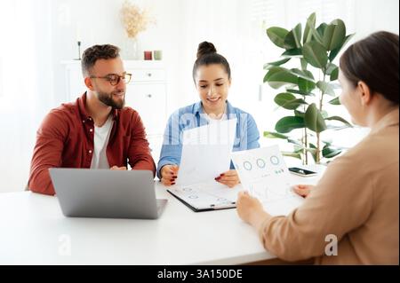 Un uomo e una donna, seduti a una scrivania in un ufficio, mostrano un grafico a un collega e lo indicano sorridente, analizzano insieme i dati aziendali, con notebook e documenti sulla scrivania Foto Stock