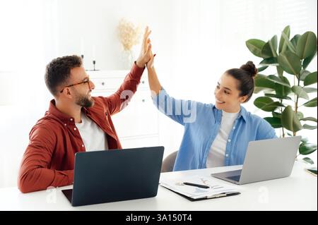 Allegri un uomo e una donna hanno cinque anni mentre sono seduti alla scrivania in un ufficio moderno, festeggiando un successo insieme, entrambi stanno lavorando sui loro laptop e si divertono a lavorare, sorridono Foto Stock