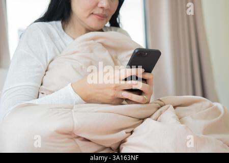 Donna asiatica anziana felice dopo il risveglio, guardando un film sdraiato sul letto in camera da letto a casa. Messa a fuoco selettiva. Foto Stock