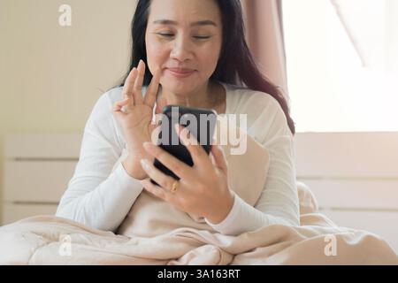 Felice donna asiatica anziana che usa il telefono cellulare dopo essersi svegliata, guardando e salutando a letto in camera da letto a casa. Concetto mattutino. Foto Stock