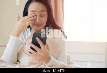 Donna matura felice in pigiama che sfrega gli occhi e usa il telefono cellulare sul letto in camera da letto. Messa a fuoco selettiva. Foto Stock