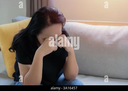 Attraente donna asiatica adulta infelice seduta sul divano nel salotto si sente triste sola e turbata per problemi di vita. Foto Stock