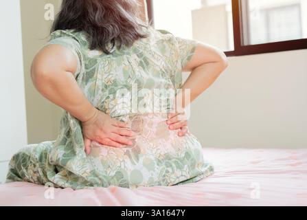 Donna anziana che soffre di mal di schiena dopo il sonno, sfregamento muscolare rigido, anziana seduta sul letto toccando la parte inferiore della schiena, sensazione di disagio. Vista posteriore, Foto Stock
