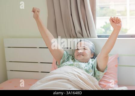 Donna asiatica anziana sdraiata sul suo letto la mattina sbadigliando con le braccia sollevate in un tratto. Salutare il nuovo giorno, concetto di buon giorno Foto Stock