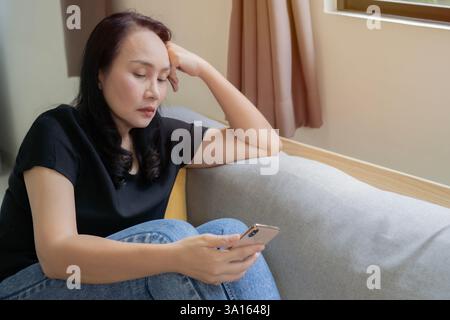 Dipendenza e depressione dallo smartphone. Donna asiatica adulta turbata seduta sul divano di casa con lo smartphone. Copia spazio. Foto Stock