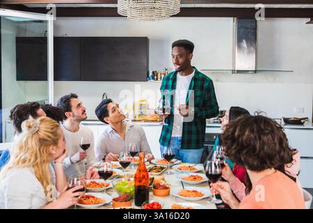 Un gruppo di amici si diverte a casa, ascoltando un giovane che fa un brindisi Foto Stock
