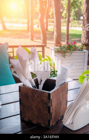 Il contenitore in legno rustico contiene posate ordinate accanto a un portatovaglioli bianco in un tranquillo giardino, invitanti cene all'aperto e relax Foto Stock