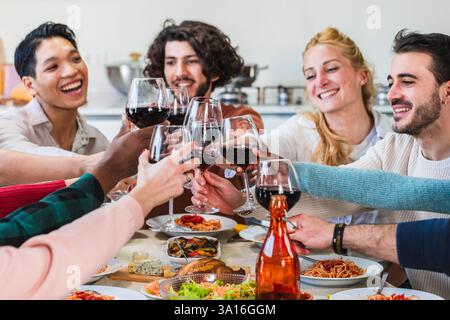 Allegri amici multietnici che brinano bicchieri di vino rosso mentre si gusta una cena italiana a casa Foto Stock