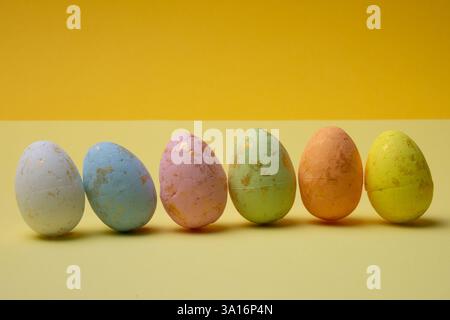 Una fila di sei uova di pasqua colorate, dipinte in tonalità pastello, si trova su uno sfondo giallo brillante. Le uova hanno piccoli accenti dorati. Foto Stock