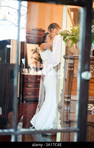 La sposa e lo sposo condividono un bacio romantico, catturato attraverso una finestra Foto Stock