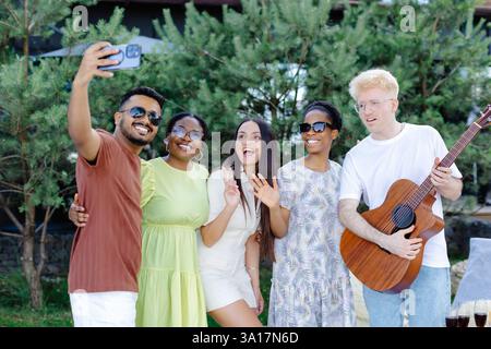 Un gruppo di amici multirazziali che si divertono e bevono vino, scattano selfie durante il picnic estivo. Foto Stock