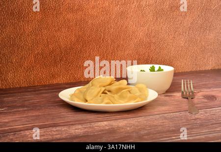 Un piatto di gnocchi appena cucinati, una forchetta e una tazza di panna acida con un rametto di prezzemolo su un tavolo di legno. Primo piano. Foto Stock