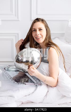 Una giovane donna in bretelle e pigiama si siede a letto tra lenzuola bianche e apre il vassoio Colazione mattutina in hotel Foto Stock