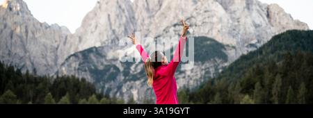Una donna con una giacca rosa solleva le braccia con gioia sullo sfondo di montagne maestose e foreste lussureggianti, abbracciando la bellezza dell'aria aperta. Foto Stock