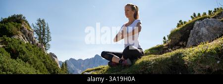 Una donna siede in una tranquilla meditazione su una collina erbosa, circondata da cime rocciose e vegetazione lussureggiante, abbracciando la tranquillità della natura. Foto Stock