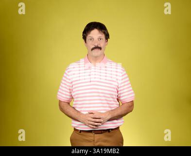 Giovane adulto dai capelli scuri con baffi, polo a righe rosa e bianca, pantaloni marroni, che guardano la macchina fotografica con un sorriso e mani agganciate in franco Foto Stock