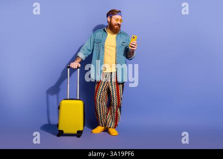 Elegante uomo barbuto in un outfit di ispirazione retrò che regge un telefono, accanto a un bagaglio giallo su uno sfondo viola Foto Stock