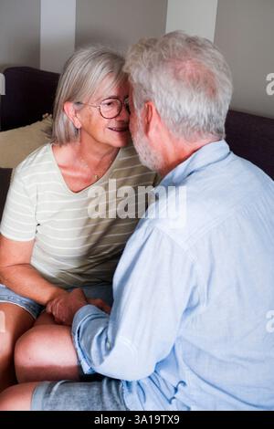 Scena emozionante di una coppia matura, un uomo e una donna amorevoli, che si coccolano sul divano di casa. I nonni condividono dolci momenti di affetto, flirtando e chiacchierando tranquillamente in casa, godendosi i loro anni d'oro insieme Foto Stock