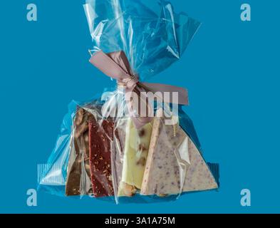 assortimento di pezzi di cioccolato in marrone, rosso, bianco, cosparso di colore in sacchetto regalo trasparente con nastro su morbido sfondo blu, spazio copia Foto Stock