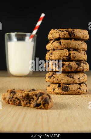 Una pila di biscotti con scaglie di cioccolato si trova su un tavolo di legno accanto a un bicchiere di latte. I biscotti sono impilati uno sopra l'altro e uno manca una torta Foto Stock