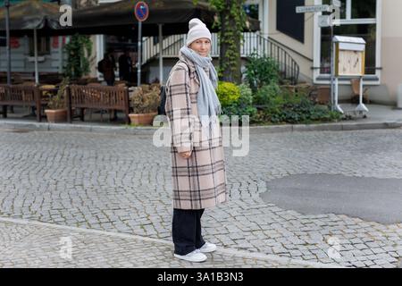 Ritratto di una donna dai vestiti caldi, indossa una sciarpa e un cappello a scacchi in una strada acciottolata a Potsdam. Confuso dubbio incerto divertente facciale Foto Stock