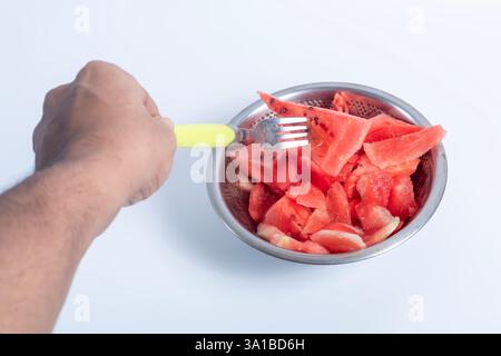 Una mano con una forchetta che si avvicina alle fette di anguria fresca poste in una ciotola in acciaio inox con sfondo bianco. Foto Stock