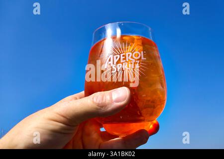 Augusta, Baviera, Germania - 7 marzo 2025: Un bicchiere di Aperol Spritz con cubetti di ghiaccio è tenuto in mano *** Ein Glas Aperol Spritz mit Eiswürfeln wird in der Hand gehalten Foto Stock
