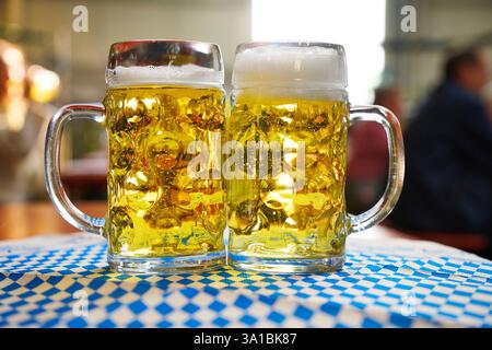 Augusta, Baviera, Germania - 7 marzo 2025: Due tazze da birra ripiene si trovano su una tovaglia bavarese *** Zwei gefüllte Bierkrüge stehen auf einer bayerischen Tischdecke Foto Stock