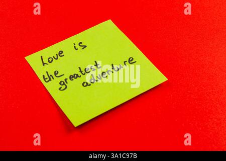 Una nota adesiva verde brillante con un messaggio scritto a mano che dice che l'amore è la più grande avventura è collocata su uno sfondo rosso brillante. I colori della nota Foto Stock