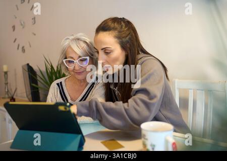 Donna che aiuta una madre anziana a utilizzare un tablet digitale a casa Foto Stock