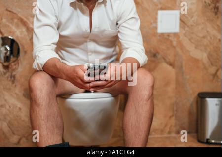 Un uomo con una camicia bianca è seduto sul gabinetto, tenendo uno smartphone tra le mani. Foto Stock