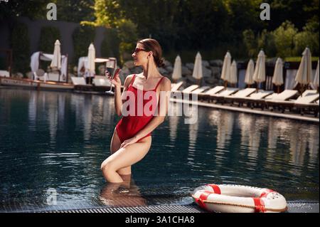 In piedi e tenendo il cocktail. La giovane donna in costume da bagno rosso è in piscina. Foto Stock
