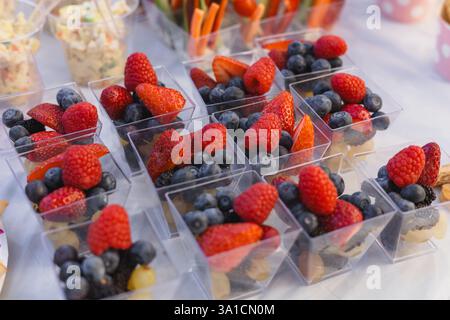 Una variegata gamma di tazze di bacche piene di fragole, mirtilli e lamponi è esposta a un raduno estivo. L'ambiente e' vivace, con decor Foto Stock