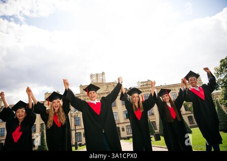 Gruppo di giovani laureati che festeggiano in berretti e abiti tradizionali di fronte a uno storico edificio universitario sotto un cielo estivo luminoso. Foto Stock