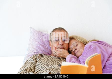 Felice coppia di anziani che si rilassa leggendo un libro in camera da letto durante il World Book Day Foto Stock